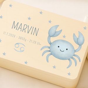 Könnte beinhalten: Eine hellfarbene Holzkiste mit einer blauen Krebs-Illustration und dem Namen "MARVIN" auf der Oberseite. Die Kiste hat auch blaue Sterne und das Krebs-Tierkreiszeichen. Ein gehäkeltes Hasenrassel und eine Holzblume befinden sich im Vordergrund.