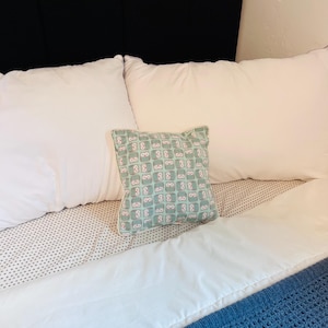 May include: A bed with white pillows and a patterned decorative pillow. The decorative pillow has a light green background with a repeating pattern of pink and white owl-like figures. A blue knitted blanket is at the foot of the bed.