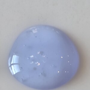May include: A circular dollop of translucent, light periwinkle blue gel with a smooth, viscous texture. The substance contains several small, suspended air bubbles throughout its body, reflecting light on its glossy, rounded surface.