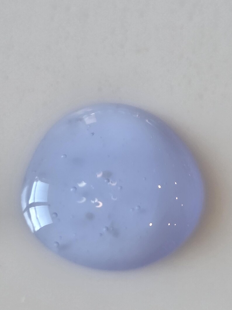 May include: A circular dollop of translucent, light periwinkle blue gel with a smooth, viscous texture. The substance contains several small, suspended air bubbles throughout its body, reflecting light on its glossy, rounded surface.
