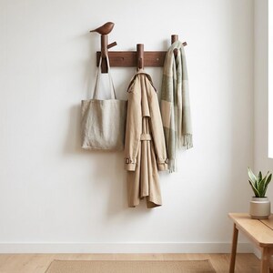 Peut inclure: Un porte-manteau mural en bois avec un ornement en forme d'oiseau. Un sac fourre-tout beige, un trench-coat beige et une écharpe verte et beige sont suspendus aux crochets. Le porte-manteau est fixé sur un mur blanc, avec un banc en bois et une plante en pot en arrière-plan.