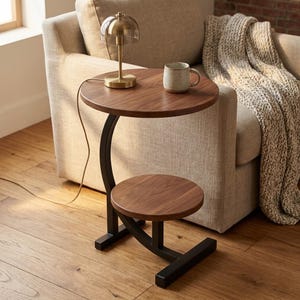 Op de afbeelding: Een bijzettafel met twee niveaus, afgewerkt met donker hout en een zwart metalen frame. De bovenste laag bevat een kleine messing lamp en een mok. De onderste laag is een kleiner, cirkelvormig oppervlak.