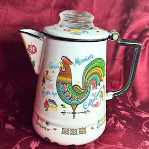 Puede incluir: Cafetera de esmalte blanco con un diseño de gallo colorido y las palabras "Good Morning" y "Coffee Time". La cafetera tiene un asa negra y una tapa de cristal. Motivos florales y de casas decoran los lados.