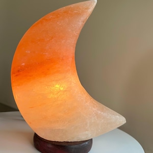May include: A crescent moon-shaped lamp carved from orange Himalayan salt, featuring a translucent, crystalline texture with warm internal illumination. The lamp is mounted on a circular, dark brown wooden base, displaying natural mineral variations.
