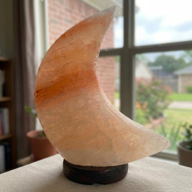 May include: A crescent moon-shaped lamp carved from translucent pink Himalayan salt, featuring natural orange and white mineral striations throughout the crystalline surface, mounted on a circular dark brown wooden base.