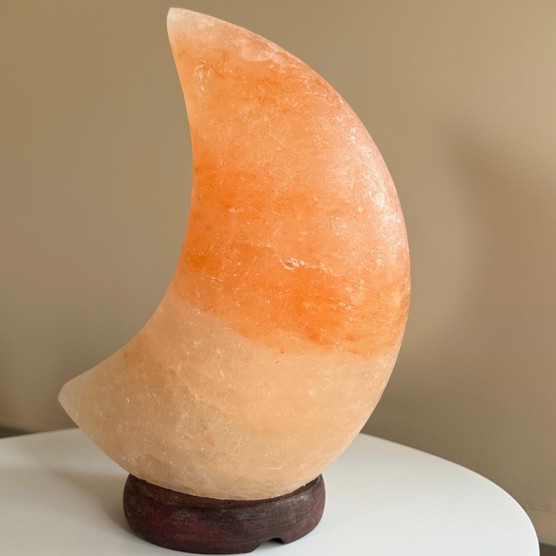 May include: A crescent moon-shaped lamp carved from natural Himalayan salt. The lamp features a gradient of translucent orange and pale peach hues with a rough, crystalline texture. It is mounted on a circular, dark brown wooden base.