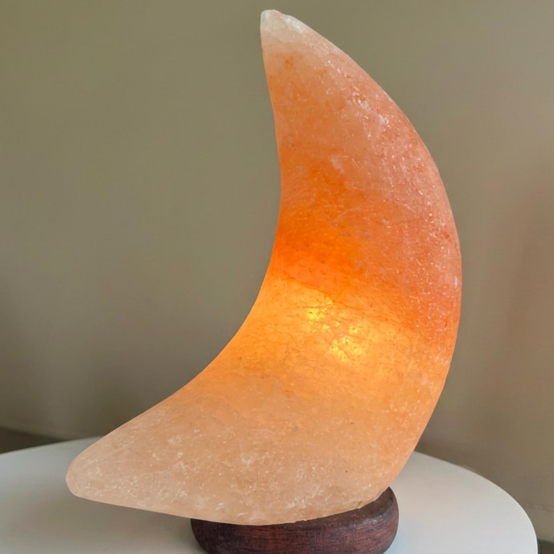 May include: A crescent moon-shaped lamp carved from translucent pink Himalayan salt. The rock salt features a textured, crystalline surface with varying shades of orange and peach. It is mounted on a circular, dark brown wooden base.
