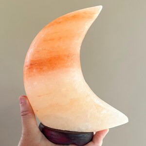 May include: A crescent moon-shaped lamp carved from translucent pink Himalayan salt, featuring natural mineral variations in shades of peach and orange, mounted on a dark, circular wooden base with a smooth, polished finish.