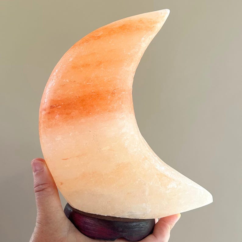 May include: A crescent moon-shaped lamp carved from translucent pink Himalayan salt, featuring natural mineral variations in shades of peach and orange, mounted on a dark, circular wooden base with a smooth, polished finish.