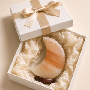 May include: A crescent moon-shaped salt lamp with orange and white marbled patterns, resting on a dark brown wooden base inside a white gift box. The box is lined with cream-colored tissue paper and topped with a matching satin ribbon bow.