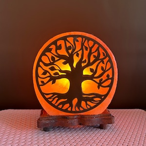 May include: A round, illuminated salt lamp with a tree of life design. The lamp emits a warm orange glow, and the tree silhouette is dark brown. It sits on a wooden base, creating a decorative and ambient light source.
