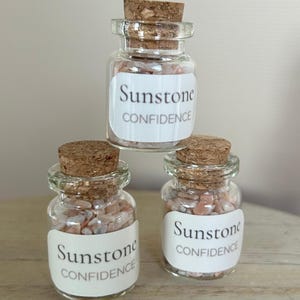 May include: Three small clear glass jars with cork stoppers, each filled with tiny, light-colored sunstone chips. Each jar features a white rectangular label with the text Sunstone CONFIDENCE printed in a dark gray serif and sans-serif font.
