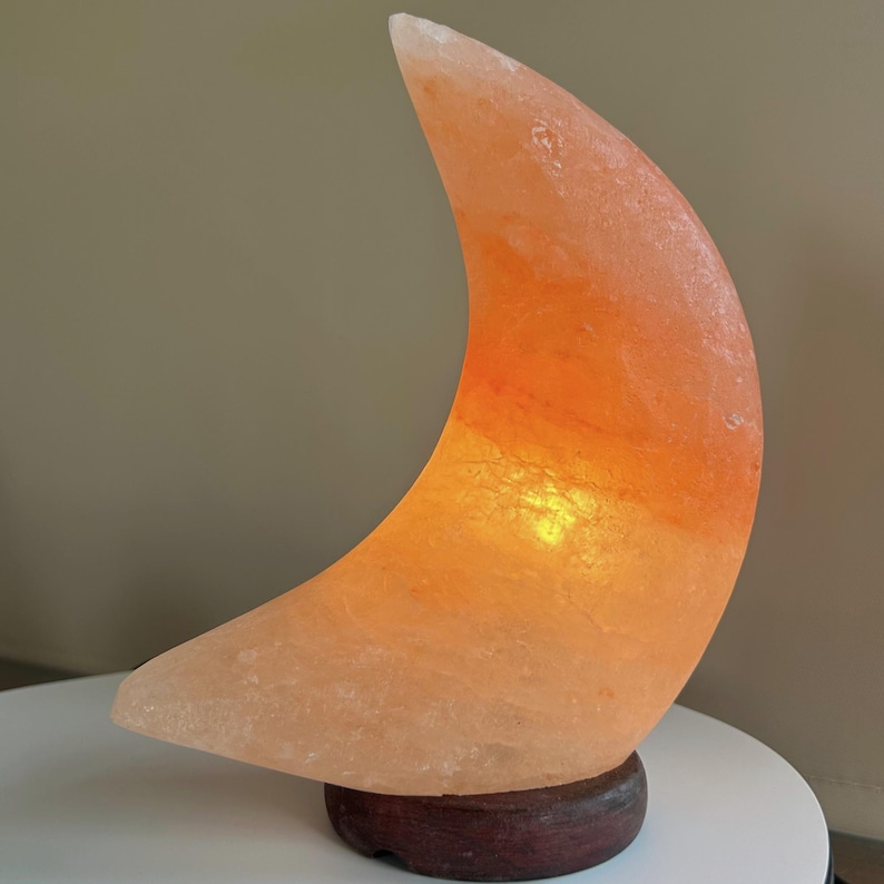 May include: A crescent moon-shaped lamp carved from natural Himalayan salt, featuring a translucent orange and pink crystalline texture. The lamp is illuminated from within, casting a warm glow, and is mounted on a circular dark brown wooden base.