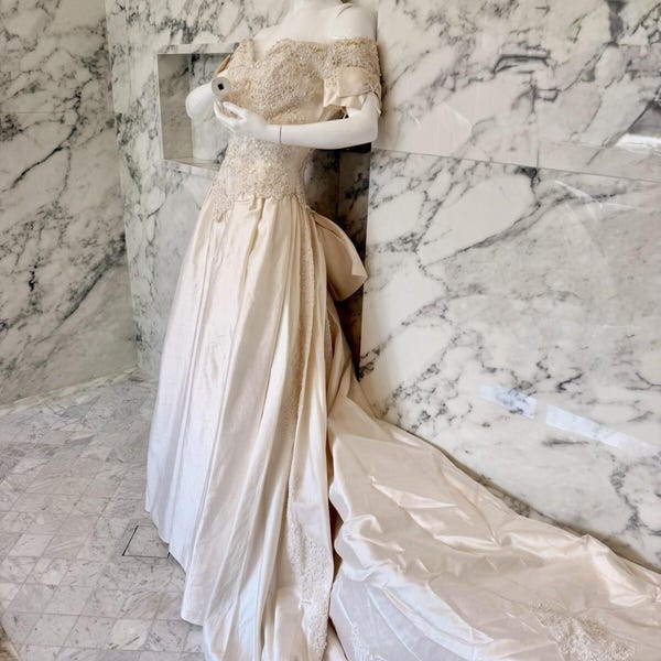 May include: An off-the-shoulder cream-colored gown featuring a fitted bodice with intricate beadwork and lace detailing. The dress has a full-length skirt with a long train, accented by lace appliques along the side and hem, and a large bow at the back.