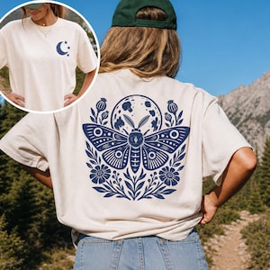 May include: An off-white t-shirt featuring a large dark blue graphic on the back of a moth, moon, and floral elements. The front chest area displays a smaller dark blue graphic of a crescent moon and stars. The shirt is worn with blue denim shorts.