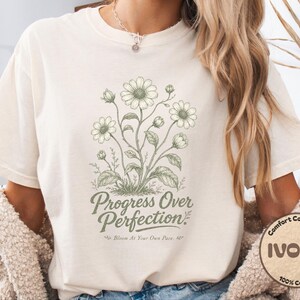 Peut inclure: T-shirt ivoire avec un motif floral et le texte "Progress Over Perfection". Le motif présente une illustration botanique de marguerites et de feuilles en vert clair. Le t-shirt est en 100% coton.