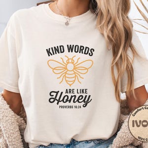 Peut inclure: T-shirt ivoire avec l'inscription "Kind Words Are Like Honey" et un graphisme d'abeille noir et or. La phrase "Proverbes 16:24" est en dessous du mot "Honey". Le t-shirt est en 100% coton.