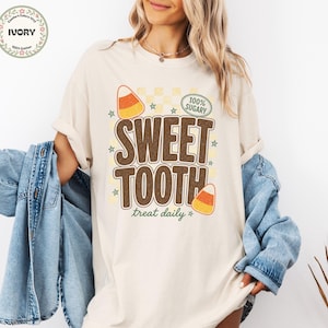 May include: An ivory short-sleeve t-shirt featuring a graphic print with the text "SWEET TOOTH" in large brown letters, "treat daily" in green script, and "100% SUGARY" in a green oval, accented by candy corn illustrations and small stars.