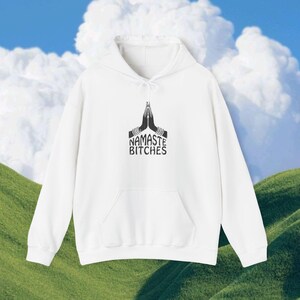 Puede incluir: Sudadera blanca con bolsillo delantero y cordón. La sudadera presenta un gráfico negro de manos en posición de oración con el texto "NAMASTE BITCHES" debajo. El fondo es un cielo con nubes y una colina verde.