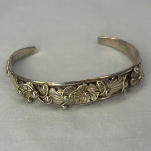 May include: Silver-toned cuff bracelet with an open design. The bracelet features a detailed floral and leaf pattern along the front. The design includes stylized flowers, leaves, and small beaded accents. The bracelet is likely made of metal.