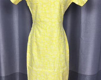 May include: A yellow dress with a high collar and short sleeves. The dress has a textured floral pattern and a fitted silhouette. The dress is displayed on a mannequin against a gray curtain backdrop.