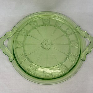 May include: A light green, round glass serving tray with two handles. The tray features a floral and geometric pattern. The center has a floral design, and the rim has a series of square floral designs. The handles are curved.