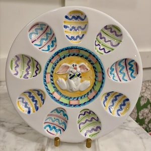 May include: White ceramic Easter egg serving platter with a central bunny design and egg-shaped indentations. The eggs are decorated with colorful wavy lines. The platter is displayed on a clear acrylic stand with gold-colored metal supports.