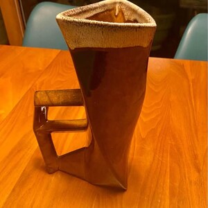 May include: A brown ceramic mug with a unique triangular shape and a handle. The top rim has a light tan color, and the body is a darker brown. The mug is on a wooden table.