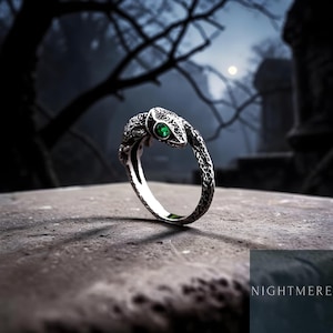 May include: A silver ring shaped like a coiled snake with a textured, dark finish and a single small, round, vibrant green gemstone set as the eye. The word NIGHTMERE is printed in white text in the bottom right corner of the image.