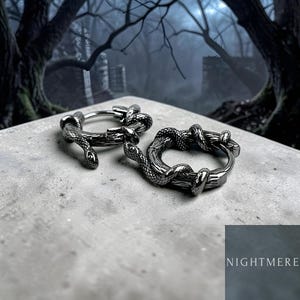 May include: A pair of silver hoop earrings with a snake and branch design. The earrings are set against a grey surface, with a blurred forest backdrop. The word "NIGHTMERE" is displayed in the bottom right corner of the image.