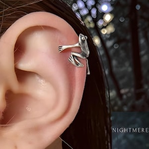 May include: A silver frog ear cuff. The frog is detailed with outstretched arms and legs, appearing to cling to the ear. The cuff is designed to wrap around the ear's cartilage. The background is blurred, with a dark, forest-like aesthetic.