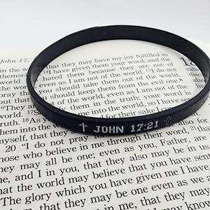 Op de afbeelding: Zwarte rubberen armband met de tekst "JOHN 17:21" en een kruis. De armband ligt op een open boek. Een religieus accessoire.