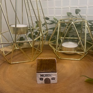 Puede incluir: Una pequeña casa de cerámica hecha a mano con un tejado marrón y la palabra "MUSIC" en la parte delantera. La casa está sobre una superficie de madera, acompañada de dos candelabros geométricos dorados con velas de té encendidas.