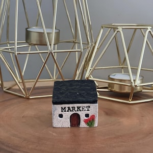 Peut inclure: Une miniature de bâtiment de marché en céramique avec un toit noir, une porte marron et le mot "MARKET" au-dessus. Des accents floraux rouges et verts sont sur le côté. Deux bougeoirs géométriques dorés sont en arrière-plan.