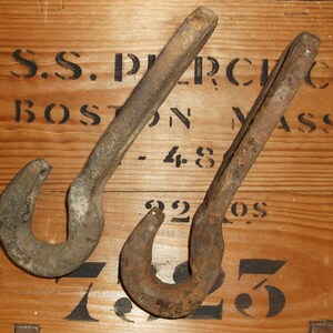 K&ouml;nnte beinhalten: Zwei rostige Metallhaken mit abgerundeter Hakenform. Die Haken befinden sich auf einer Holzoberfl&auml;che mit schwarzem Text, der "S.S. PERCH CO. BOSTON MASS. - 48 3205" lautet.