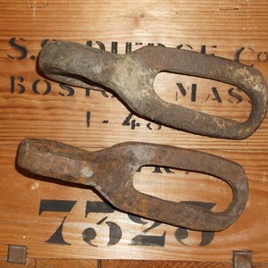 K&ouml;nnte beinhalten: Zwei rostige Metallringe mit abgerundeter Kante und ovaler &Ouml;ffnung. Die Ringe liegen auf einer Holzoberfl&auml;che mit schwarzem Text, der "S.G. PIERCE CO. BOSTON, MASS. 1-48 7529" lautet.