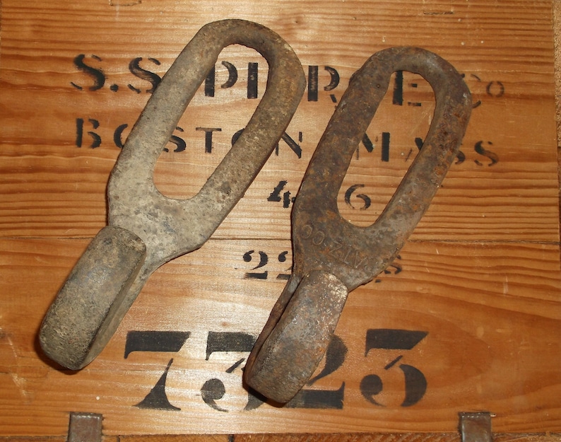 K&ouml;nnte beinhalten: Zwei rostige Metallhaken mit abgerundeter Oberseite und spitzer Unterseite. Die Haken liegen auf einer Holzoberfl&auml;che mit schwarzen Zahlen und Buchstaben. Die Buchstaben lauten "S.S. PIERCE CO BOSTON MASS 46 22 725".