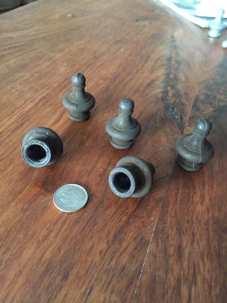 May include: A collection of six antique metal finials, with a dark, aged finish. The finials have a decorative, rounded top and a threaded base. A U.S. dime is included for size reference. The objects are arranged on a wooden surface.