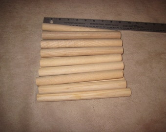 May include: A stack of 10 smooth, unfinished wooden dowels, approximately 12 inches long, arranged in a neat pile on a brown surface.