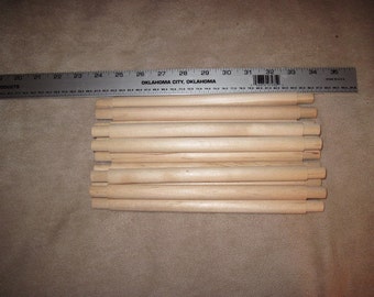May include: A stack of 15 wooden dowels, approximately 12 inches long, arranged in a staggered pattern. The dowels are unpainted and have a natural wood finish. A ruler is visible in the background, showing the length of the dowels.