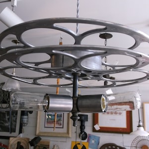 May include: A silver film reel chandelier with three clear glass light bulbs. The chandelier is suspended from the ceiling with a black wire.