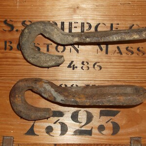 K&ouml;nnte beinhalten: Zwei rostige Metallhaken mit einer abgerundeten Hakenform. Die Haken befinden sich auf einer Holzoberfl&auml;che mit schwarzem Text, der "S.S. PIERCE CO. BOSTON MASS 486 625" lautet.