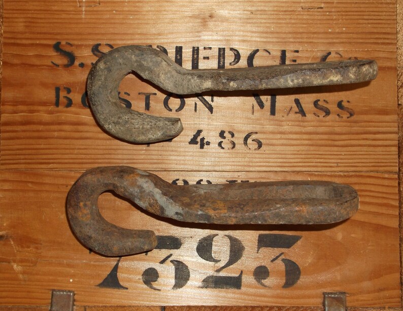 K&ouml;nnte beinhalten: Zwei rostige Metallhaken mit einer abgerundeten Hakenform. Die Haken befinden sich auf einer Holzoberfl&auml;che mit schwarzem Text, der "S.S. PIERCE CO. BOSTON MASS 486 625" lautet.
