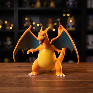 Könnte beinhalten: Eine leuchtend orangefarbene Charizard-Figur mit ausgebreiteten Flügeln und einem flammenden Schwanz. Die Figur hat ein detailliertes Design mit blaugrünen Flügeln und weißen Krallen. Sie steht auf einer Holzoberfläche, mit einem verschwommenen Hintergrund aus Regalen und Dekorationsartikeln.