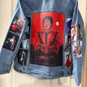Könnte beinhalten: Hellblaue Jeansjacke mit einer großen roten Grafik von Michael Jackson aus dem Musikvideo "Thriller" auf dem Rücken. Die Jacke hat mehrere kleinere Bilder von Michael Jackson auf den Ärmeln. Das Wort "Thriller" ist auf der Rückseite aufgedruckt.
