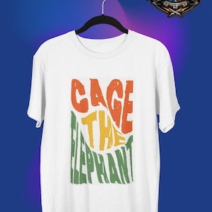 Könnte beinhalten: Weißes T-Shirt mit farbenfrohem Grafikdesign. Das Design zeigt die Worte "CAGE THE ELEPHANT" in orangefarbenen, gelben und grünen Blockbuchstaben. Das Shirt hängt an einem schwarzen Kleiderbügel vor blauem Hintergrund.