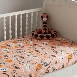May include: A white wooden crib featuring a fitted sheet with a light peach background and a repeating pattern of black and orange celestial, mystical, and skeletal motifs. A coiled snake plush toy with a black and orange diamond pattern sits on top.