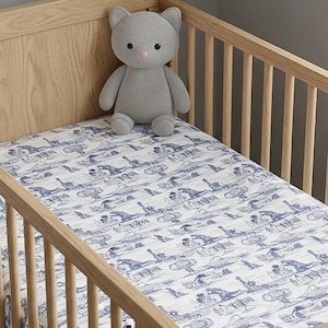 May include: A light wood crib with vertical slats and a fitted sheet featuring a blue and white toile pattern depicting various scenes. A plush gray cat-shaped stuffed animal sits upright on the mattress against the back panel of the crib.