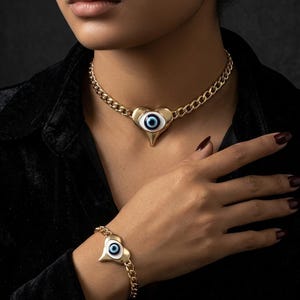 May include: A gold-tone chain necklace and matching bracelet set, each featuring a central heart-shaped pendant with a white, blue, and black evil eye motif. The jewelry pieces are constructed with thick, interlocking curb chain links.