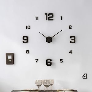 May include: A contemporary black wall clock featuring oversized numerals and hands. The clock is affixed to a white wall, accompanied by a small, dark electronic device on the left. The clock's numbers are approximately 30 cm in height.